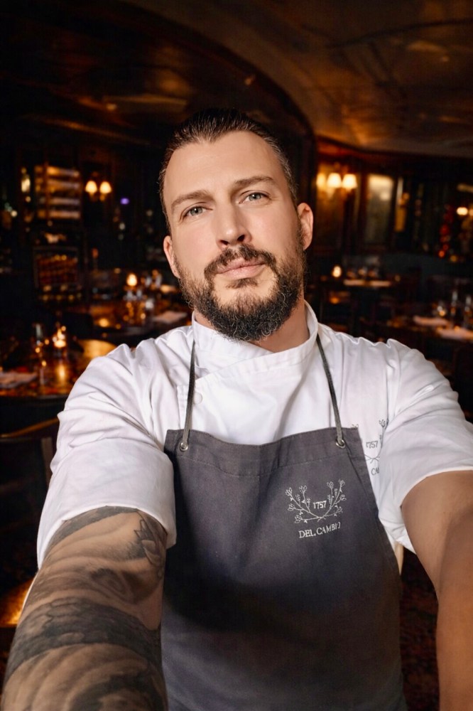 Chef with tattoos in a restaurant, wearing a white coat and dark apron, taking a selfie.
