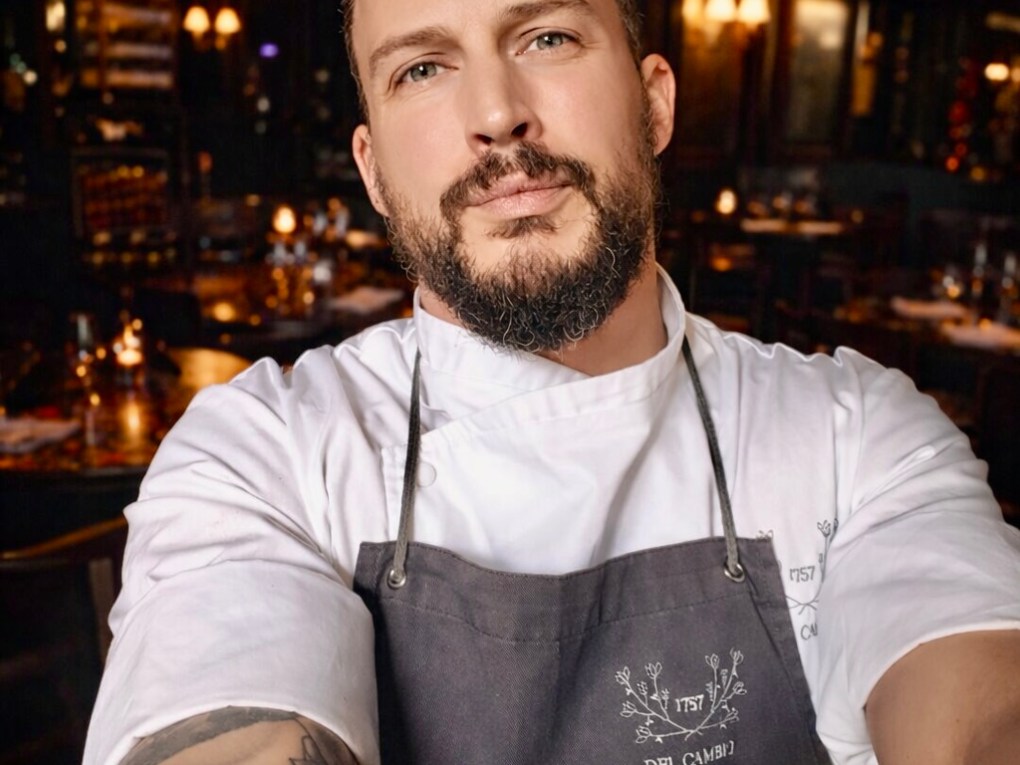 Chef with tattoos in a restaurant, wearing a white coat and dark apron, taking a selfie.