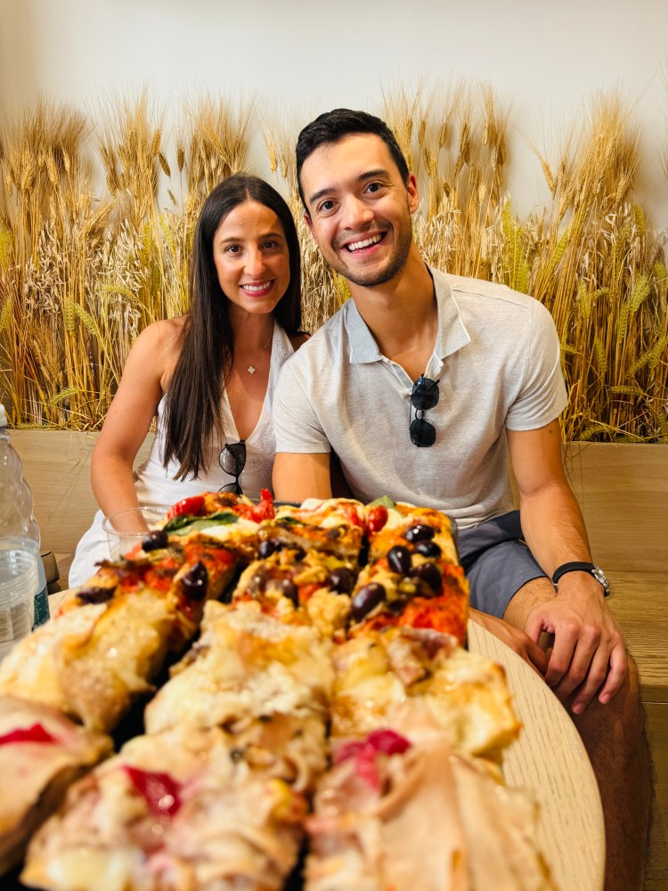 Smiling couple seated behind a plate of assorted sliced pizza.