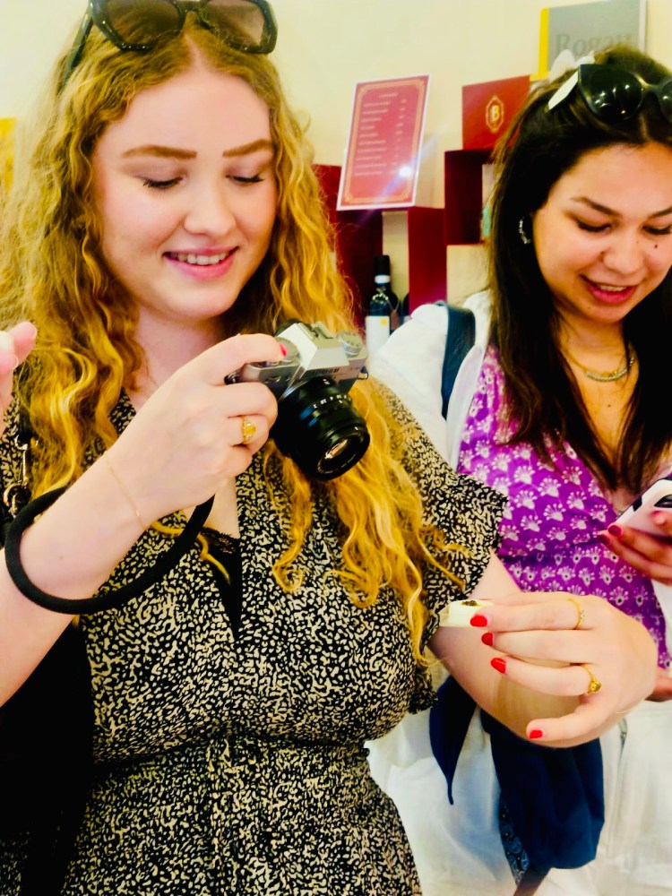Two women smiling, holding a camera and a phone indoors.