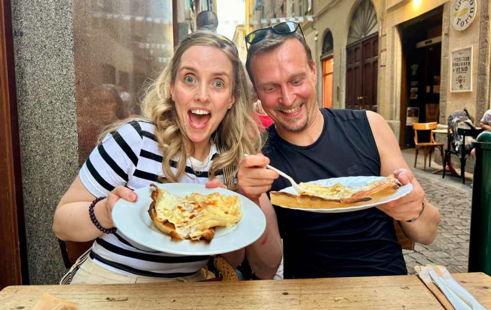 Smiling couple holding plates of food, seated at an outdoor café.