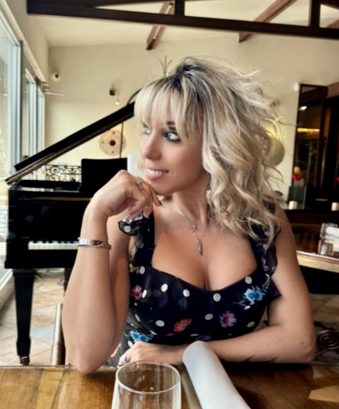 Woman with blond hair in polka dot dress sitting in front of a piano.