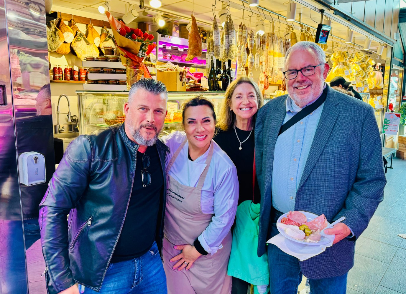 Four people smiling in a market with hanging meats and shelves of goods.
