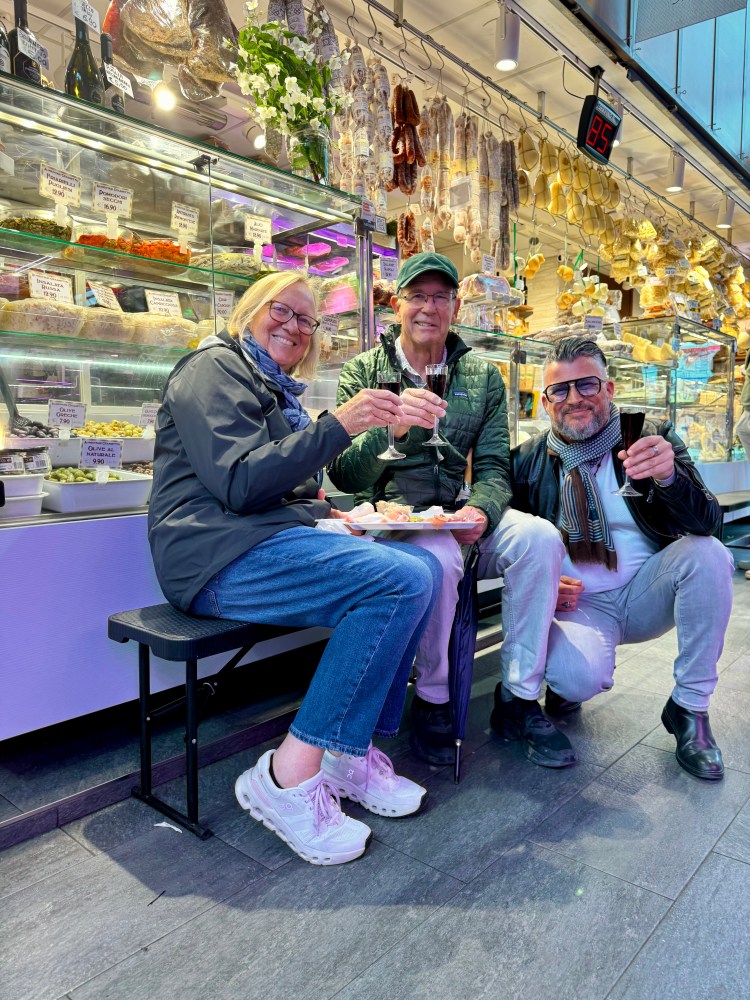 Three people seated, enjoying snacks and drinks in a market with deli items behind them.