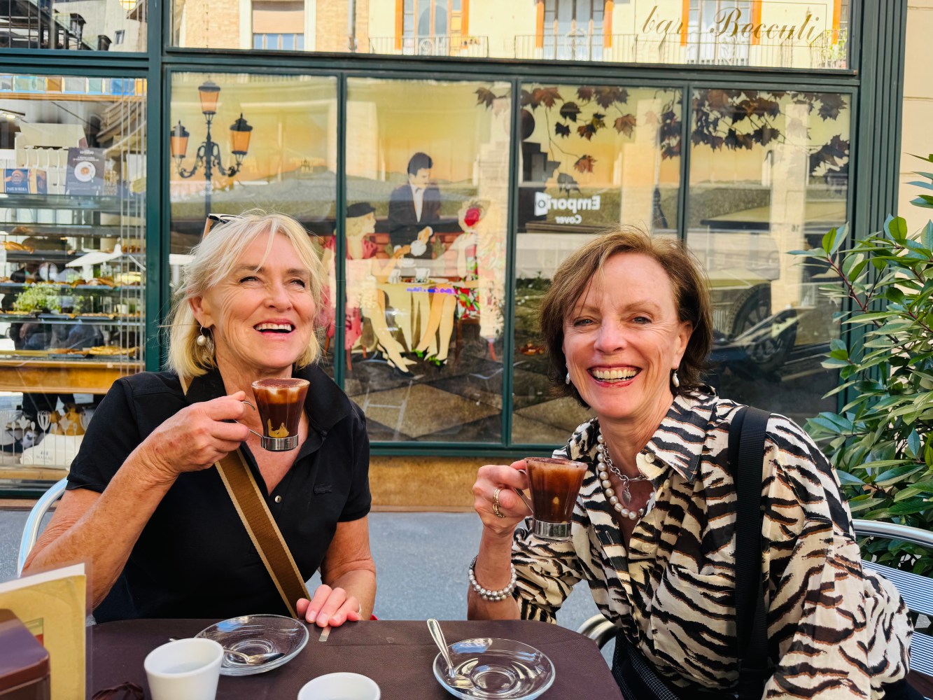 Two women smiling and holding espresso cups at a café table outdoors.